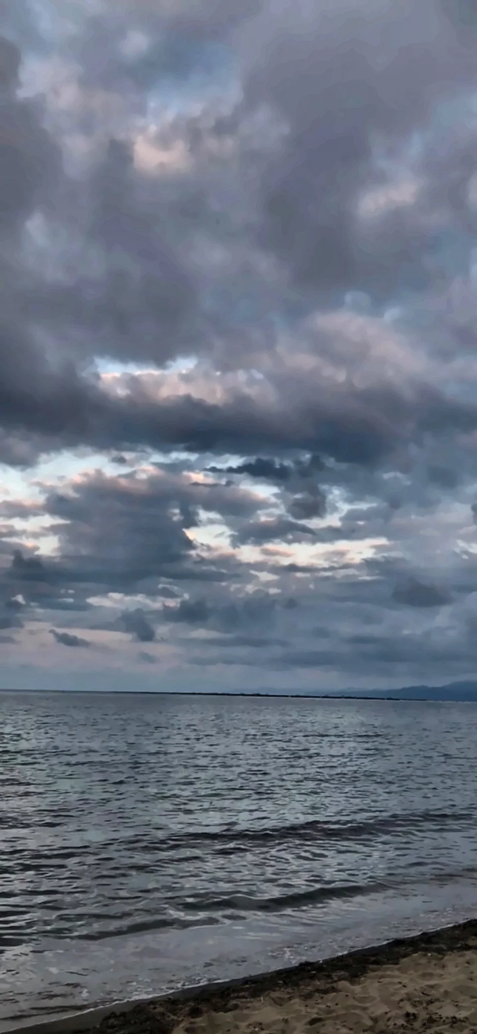 Gri bulutların altında dalgalı deniz ve çakıllı kıyı.
