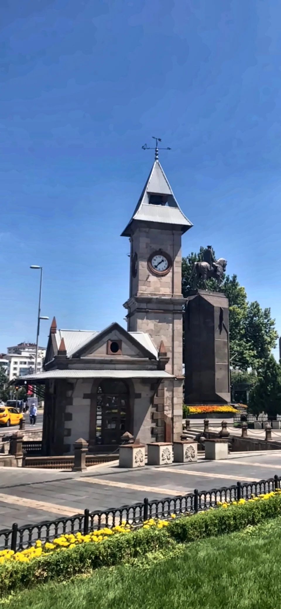 Mavi gökyüzü altında bir saat kulesi ve çevresindeki meydan.